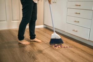 Person sweeping wooden kitchen floor with a broom, showing daily cleaning routine.
